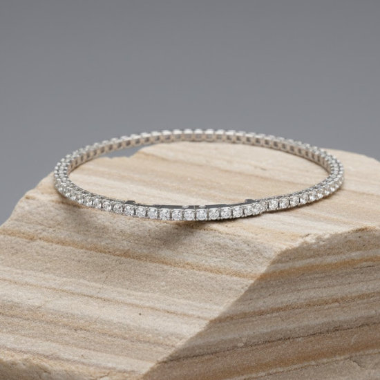 A delicate white gold diamond tennis bracelet laid flat on a light brown, layered stone display block against a solid gray background.