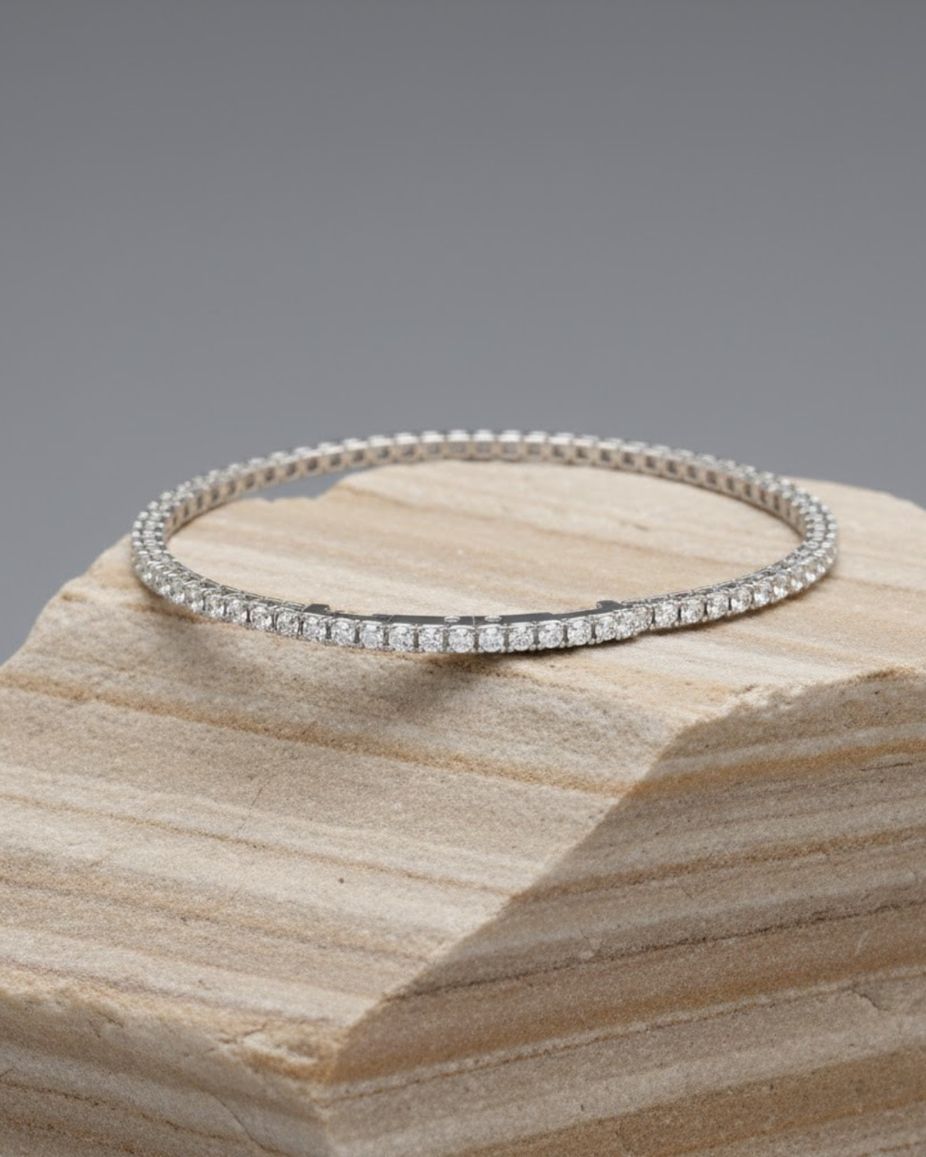 A delicate white gold diamond tennis bracelet laid flat on a light brown, layered stone display block against a solid gray background.
