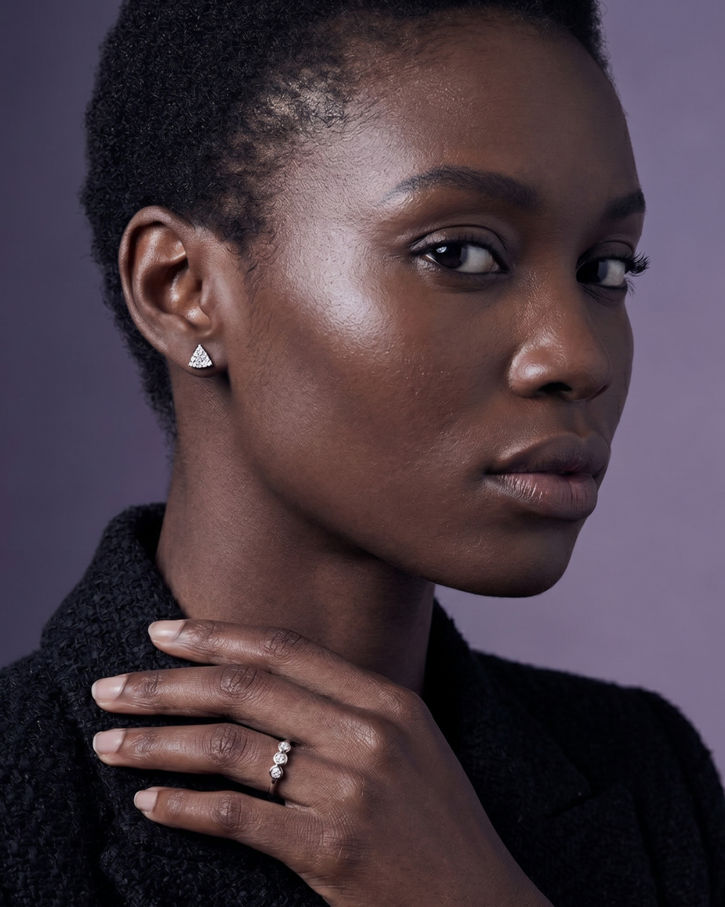 Model wearing small triangular pave diamond stud earrings and a dainty three-stone bezel ring, styled with a black tweed blazer.