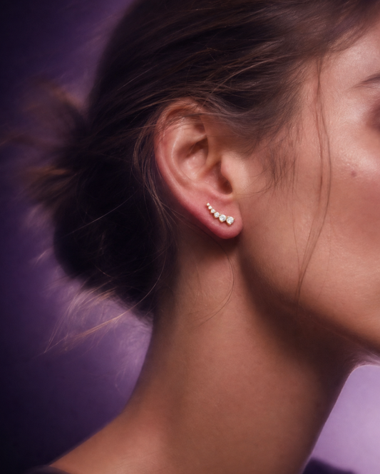Detailed close-up view of a model's ear lobe wearing a single yellow gold and diamond ear climber earring, featuring a curved sequence of multiple brilliant-cut diamonds with a distinct top cluster, set against a moody blurred purple and lavender gradient background.