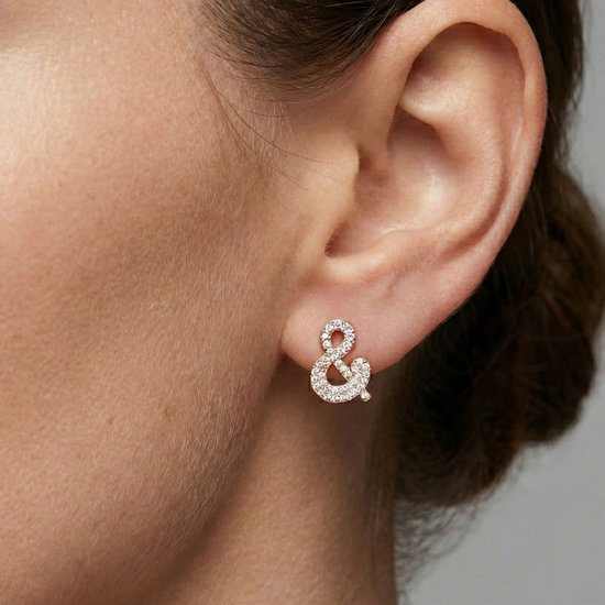 A pair of 14K yellow gold stud earrings shaped like an ampersand and covered in pavé diamonds, shown from the front on the ear.