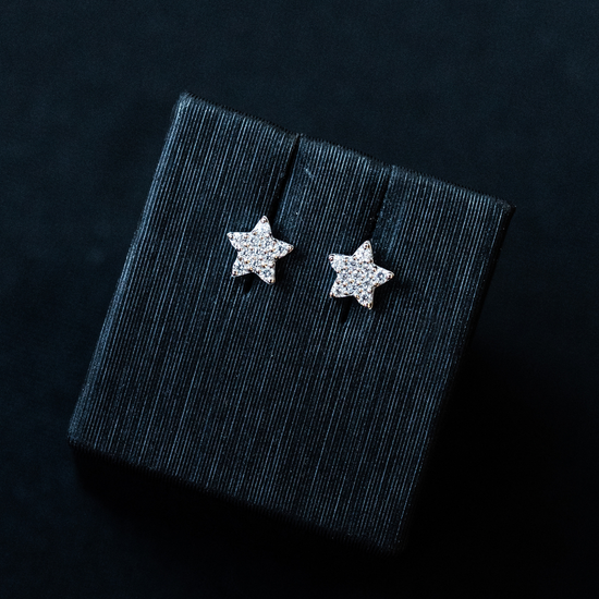A pair of sparkling, star-shaped stud earrings displayed on a dark, textured jewelry box insert against a black background.