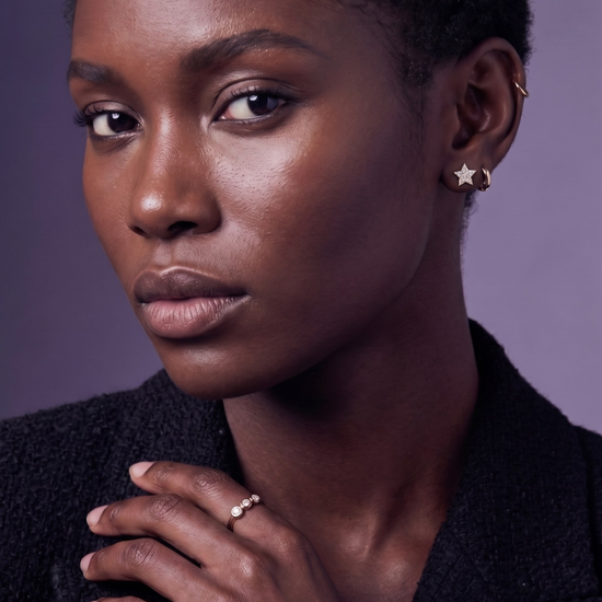 A Black woman wears a gold star diamond earring and a diamond trilogy ring against a lavender background.