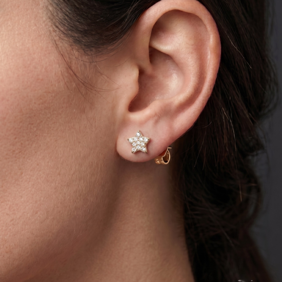 A close-up photograph of a woman's ear wearing a single gold, star-shaped stud earring paved with diamonds.