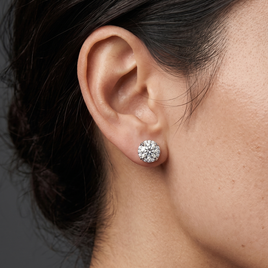 A close-up side profile shot focusing on a person's ear and jawline against a dark gray studio background. The person has dark hair pulled back and is modeling a large, sparkling round-cut diamond stud earring resting on the earlobe.