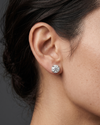 A close-up side profile shot focusing on a person's ear and jawline against a dark gray studio background. The person has dark hair pulled back and is modeling a large, sparkling round-cut diamond stud earring resting on the earlobe.