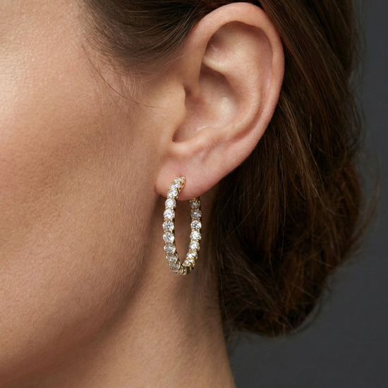 A close-up of a pair of 14K yellow gold huggie hoop earrings with a continuous line of brilliant round-cut diamonds, shown on a black background to highlight their sparkle on ear.