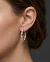 A close-up of a pair of 14K yellow gold huggie hoop earrings with a continuous line of brilliant round-cut diamonds, shown on a black background to highlight their sparkle on ear.