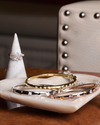 High-polish gold and silver diamond bangles arranged on a marble dish beside a diamond ring on a cone display, styled on a wooden surface with an upholstered chair in the background.