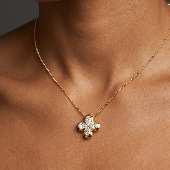 Close-up of a person's neck and collarbone wearing a delicate gold chain necklace with a small, gold, four-petal floral or clover pendant featuring diamond accents.