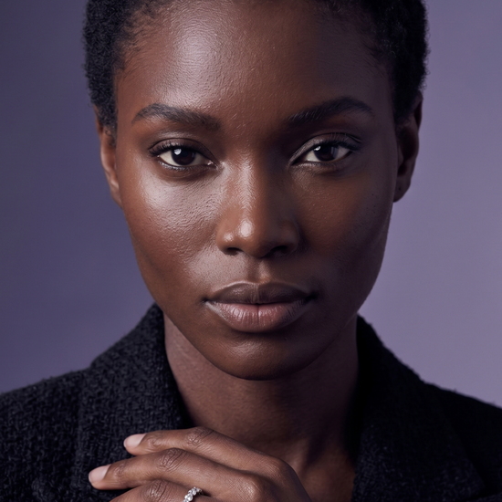 Close-up of a sparkling diamond eternity band worn by a model in a black tweed blazer against a soft violet background.