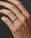 A model's hand wearing a delicate white gold or platinum half-eternity ring featuring round-cut diamonds against a dark background.
