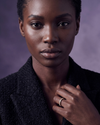 Gold diamond eternity band ring worn on the finger of a model in a black tweed blazer against a muted purple background.