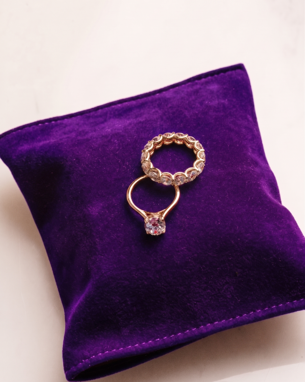 Yellow gold Amorati Bold Bezel 7-Carat Round Diamond Eternity Band displayed on a vibrant purple velvet pillow, styled with a round solitaire engagement ring.