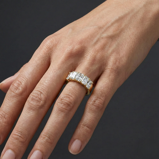A top-down view of a 14K yellow gold wide-band ring with five horizontally set emerald-cut diamonds, with the center one larger on hand.