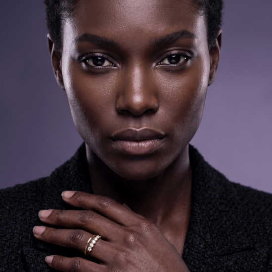 Close-up of a woman in a black tweed blazer modeling a modern gold split-shank ring featuring five diamonds against a deep violet background.
