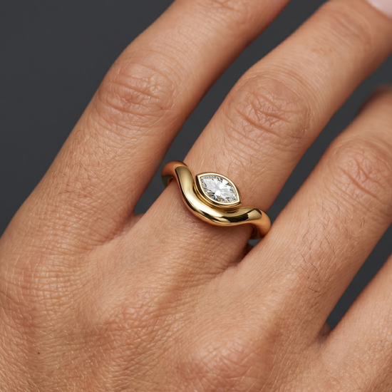 Close-up lifestyle view of a modern 14k yellow gold ring featuring a horizontally set marquise-cut diamond in a sleek bezel setting on a fluid, wavy comfort-fit band.