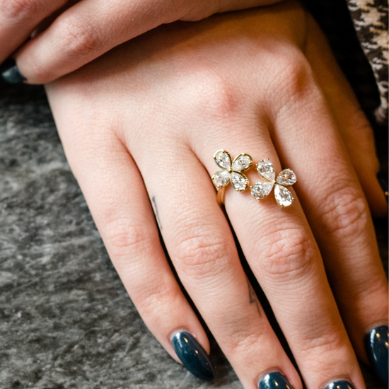 A close-up photograph of a hand with long, glossy, dark teal fingernails. On the ring finger is a yellow gold ring featuring two blooming flower motifs side-by-side, each crafted with four pear-cut diamonds. In the background, another hand is visible wearing large stacked diamond rings, including a prominent emerald-cut diamond.