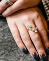 A close-up photograph of a hand with long, glossy, dark teal fingernails. On the ring finger is a yellow gold ring featuring two blooming flower motifs side-by-side, each crafted with four pear-cut diamonds. In the background, another hand is visible wearing large stacked diamond rings, including a prominent emerald-cut diamond.