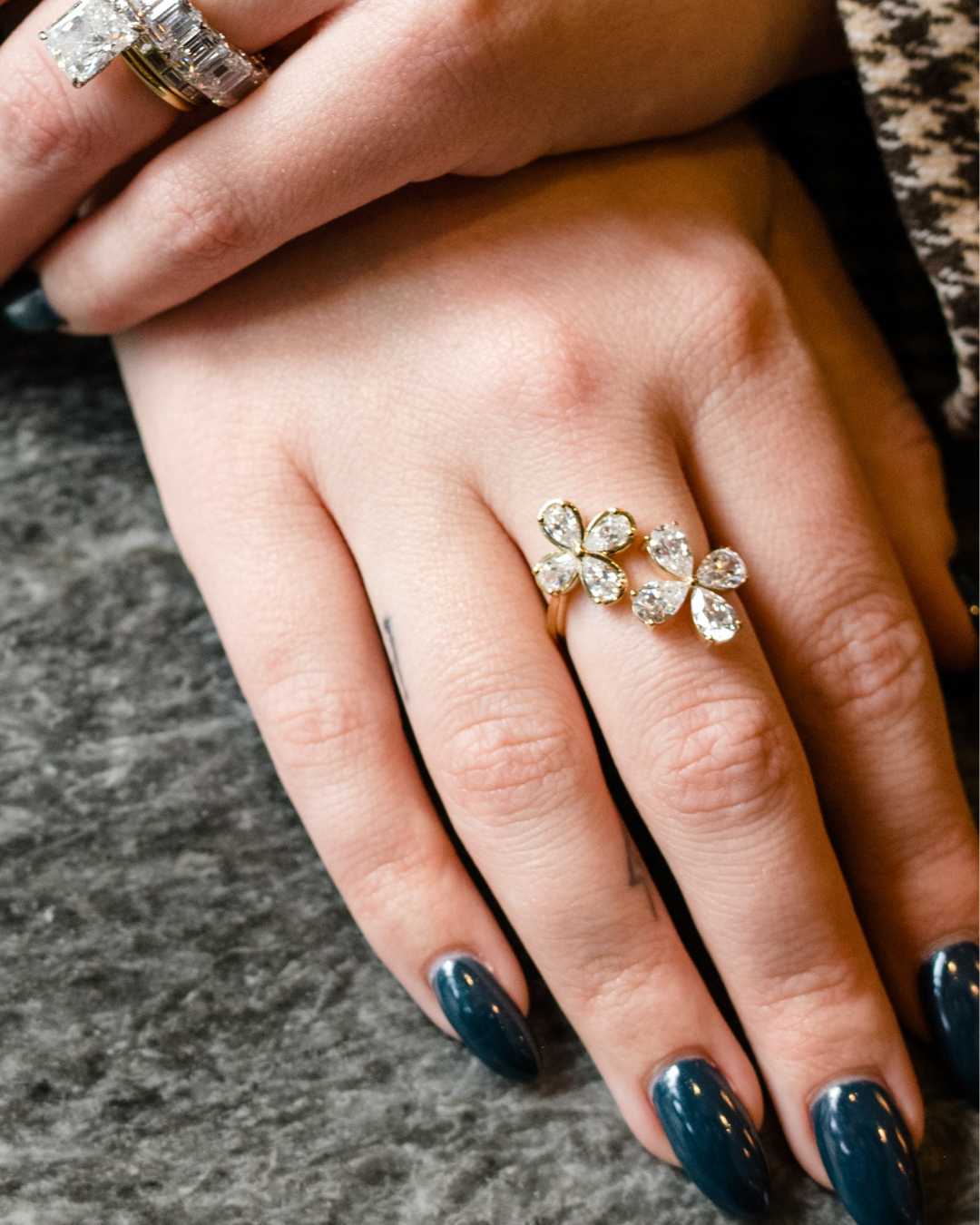 A close-up photograph of a hand with long, glossy, dark teal fingernails. On the ring finger is a yellow gold ring featuring two blooming flower motifs side-by-side, each crafted with four pear-cut diamonds. In the background, another hand is visible wearing large stacked diamond rings, including a prominent emerald-cut diamond.