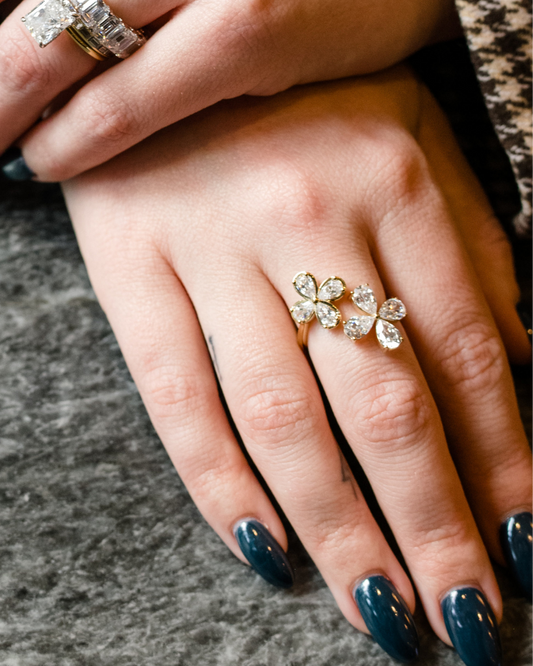 A close-up photograph of a hand with long, glossy, dark teal fingernails. On the ring finger is a yellow gold ring featuring two blooming flower motifs side-by-side, each crafted with four pear-cut diamonds. In the background, another hand is visible wearing large stacked diamond rings, including a prominent emerald-cut diamond.
