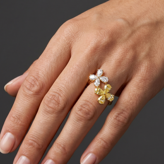 Close-up of a hand wearing a gold ring featuring two diamond flowers, one made of white diamonds and the other of yellow diamonds, against a dark gray background.