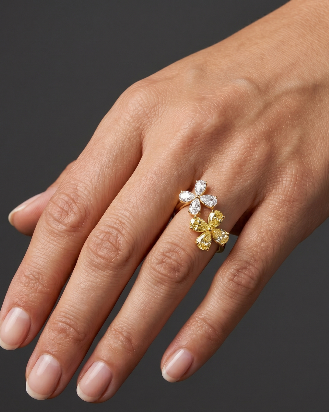 Close-up of a hand wearing a gold ring featuring two diamond flowers, one made of white diamonds and the other of yellow diamonds, against a dark gray background.